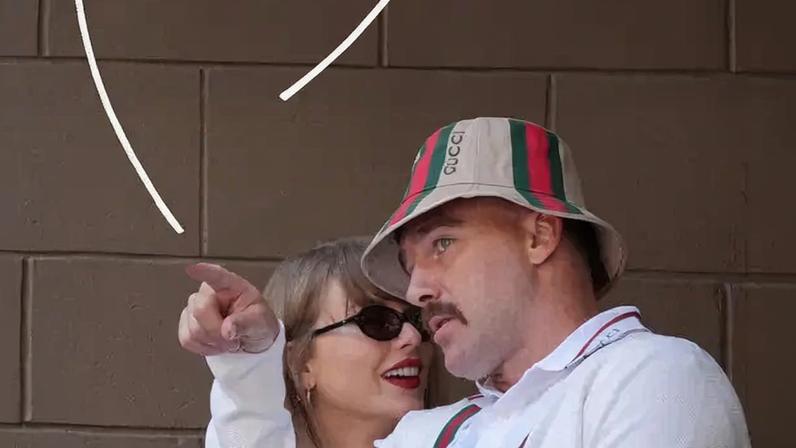 Taylor Swift, left, wearing sunglasses and red lipstick, stands behind Travis Kelce and rests her head on his shoulder. He is wearing a Gucci hat, and white Gucci polo shirt and sweater, as he draws a white heart with his forefinger in the photo. A gray cinderblock wall is behind them.