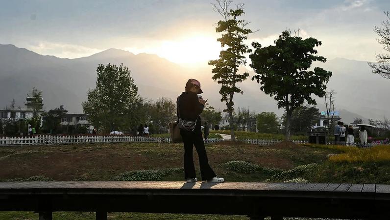 A young woman standing on a boardwalk is seen from behind as she looks down at her phone with a beautiful backdrop of mountains, trees, and open parkland beyond her.