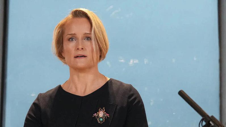 A woman in a black top with a large broach against a blue background.