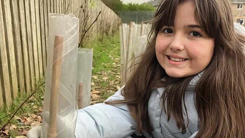 Gabriella, aged 10, wears a turquoise coat and holds a tree planter wrapped in clear plastic. There a several planters behind her. She sits crouched in a green garden. She has long brown hair.