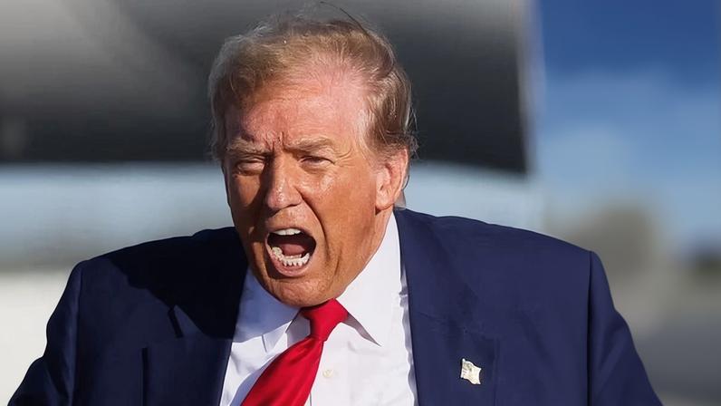 In a blue suit and red tie, Donald Trump stands with his mouth open, suggesting that he is shouting
