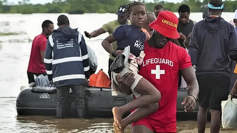 EPA/Shutterstock A man in a red T-shirt and hat with the word 'Lifeguard' on helps a girl through flood waters in southern Mozambique. Behind them is a rubber speed boat with men around it.