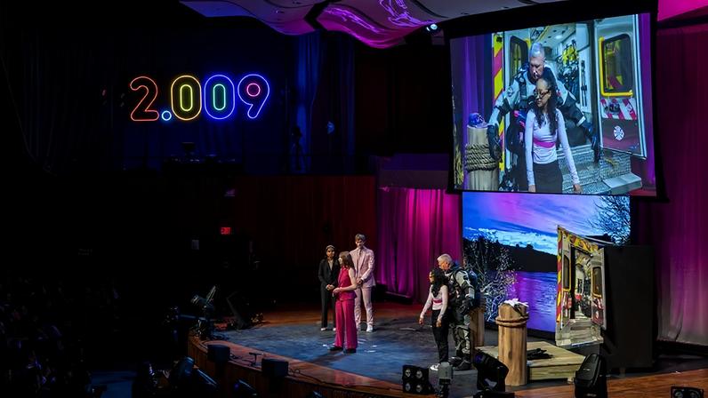 Wide shot of a stage. Two presenters demonstrate a device designed for rescue divers while three others look on.