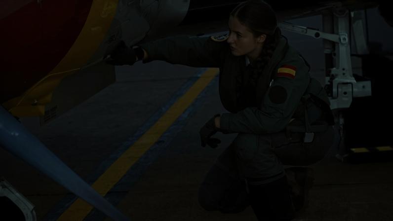 Princess Leonor crouches down under the wing and touches the fuselage of a light aircraft; she is wearing khaki overalls with the Spanish flag on the shoulder, gloves and boots, and has long, fair hair in a thick plait.