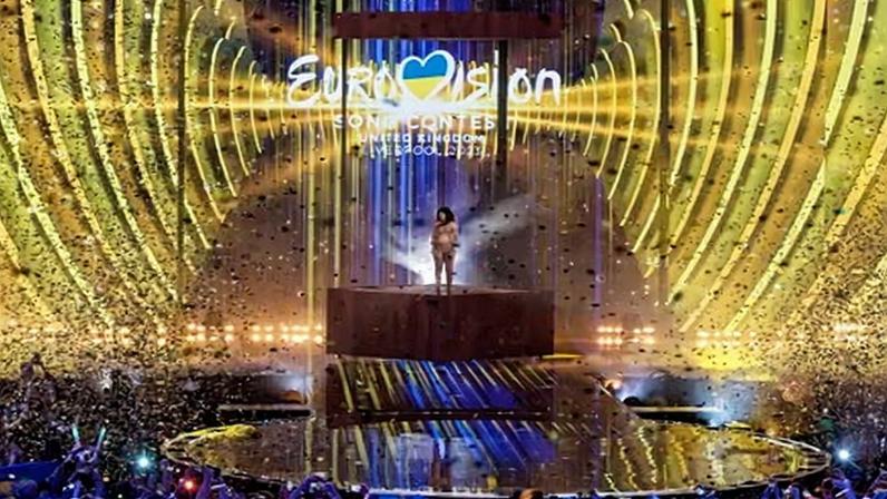 Loreen stands on a raised podium on stage under Eurovision branding as the crowd looks on