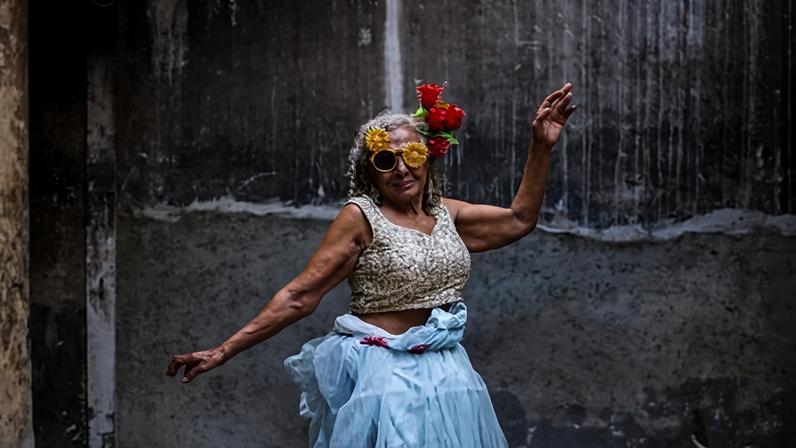A woman in her seventies holds a ballerina-like pose wearing a blue tulle skirt and a red flower in her hair.