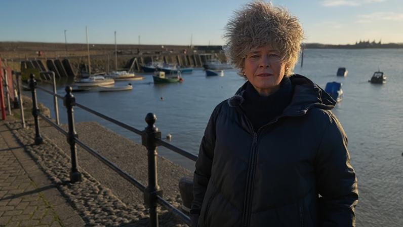 Rachel Gilmour MP is standing by Minehead harbour wearing a coat and fluffy hat. There are small boats in the harbour behind her. She is standing in front of a railing.