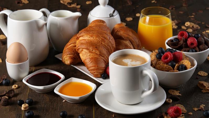 Café da manhã composto com croissant, café, suco de laranja, ovo poché, granola e leite sobre a mesa rústica de madeira.