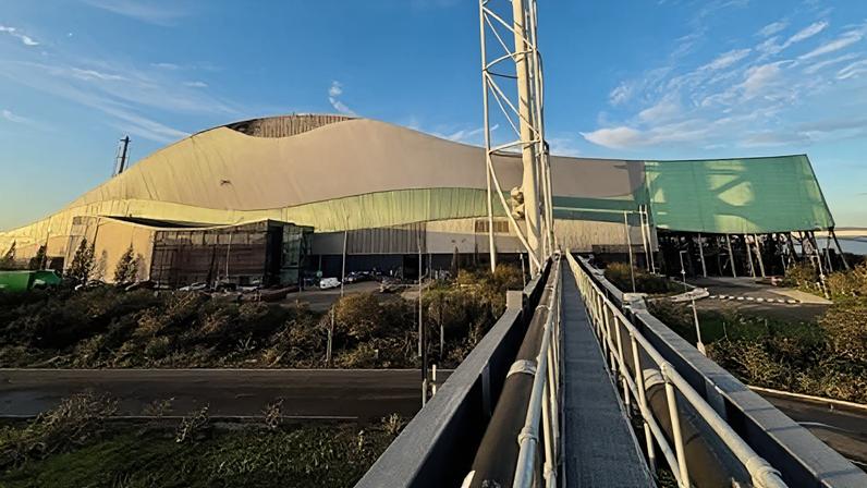 A large, modern building with a distinctive wave-like roof design, combining metallic and green-tinted panels. The structure appears industrial or possibly a sports or event venue, with a sleek, contemporary style. In the foreground, a metal walkway with railings extends toward the building, creating strong leading lines. To the right of the walkway, a tall, white structural tower rises vertically, adding to the industrial aesthetic. The surrounding area includes a road, landscaped greenery, and several parked vehicles near the entrance. The sky above is clear with scattered clouds, bathed in warm sunlight, giving the scene a bright and crisp appearance.
