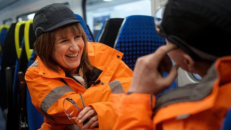 Reeves seated onboard train in orange hi-vis and hat opposite another person