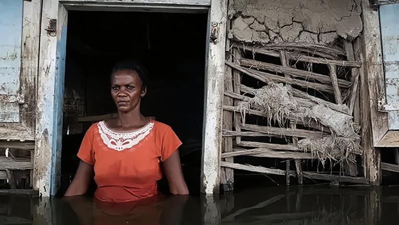 Getty Images A woman in an orange dress stands in the doorway of her flooded house on the outskirts of the city of Gonaives in Haiti. The water level is at waist height. The walls of the house are substantially damaged.