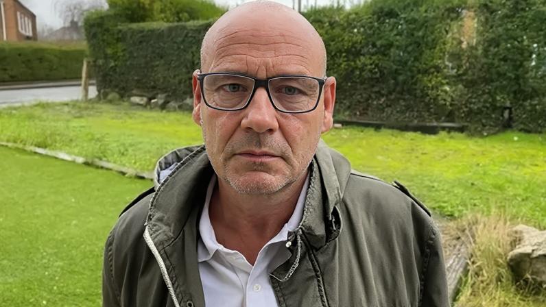 Barry Froggatt in a picture. He is standing on a front drive looking at the camera. He is making a serious face. He is a bald man wearing glasses, a white polo top and a green jacket.
