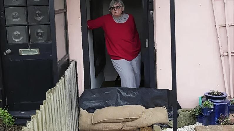 Sue Allen standing in the doorway of her pink-coloured home. She is wearing a red jumper and glasses. She is standing in front a pile of sandbags.