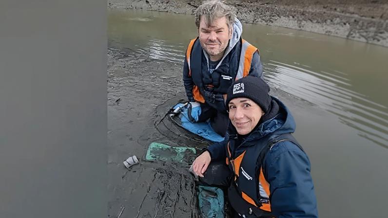 Dr Martina Bristow and engagement officer Blair Watson take a selfie during seagrass planting at the River Don near Jarrow. They are wearing high visibility blue and orange vests as they sit in the mud next to a frame which holds the young seagrass plants. Dr Bristow has brown eyes and a pierced nose and lip. She is wearing a black beanie. Blair Watson has short grey hair and blue eyes.