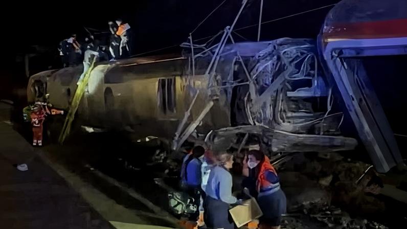 Rescue workers beside and on top of a derailed train on its side with a crumpled end, at night