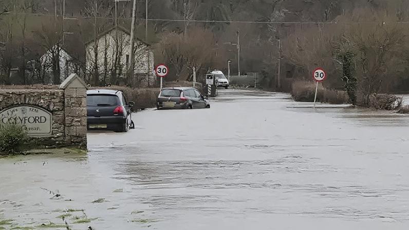 Jordan Langridge Two cars are submerged in floodwater on a road in Colyford, Devon. A sign with the village's name on it attached to a brick wall is in the foreground. A pair of 30mph signs are in the distance.