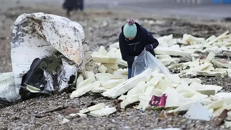 Eddie Mitchell A person putting white blocks of foam into a large, clear plastic bag on a pebble beach