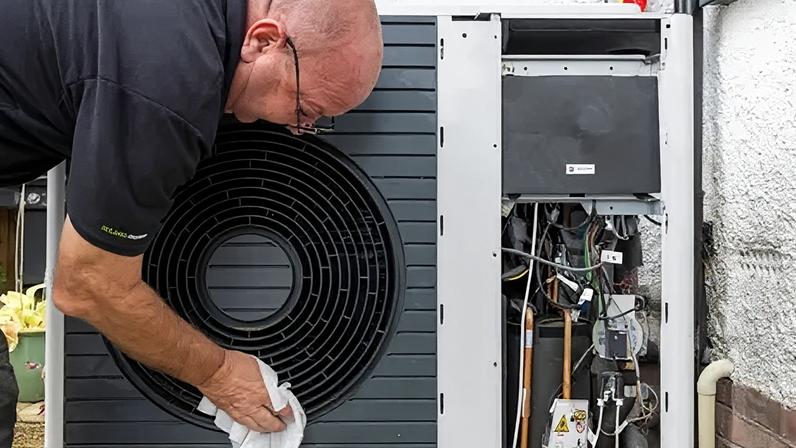 Andrew Aitchison/Getty Images An engineer - a man wearing a black T-shirt - wipes the outside of a black heat pump with a cloth. To the right of the picture the heat pump abuts the wall of a property with the pipes and electrical components visible