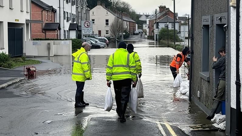 BBC Riverside area road is flooded, people in high-vis carry sandbags to doors and by the river bridge.