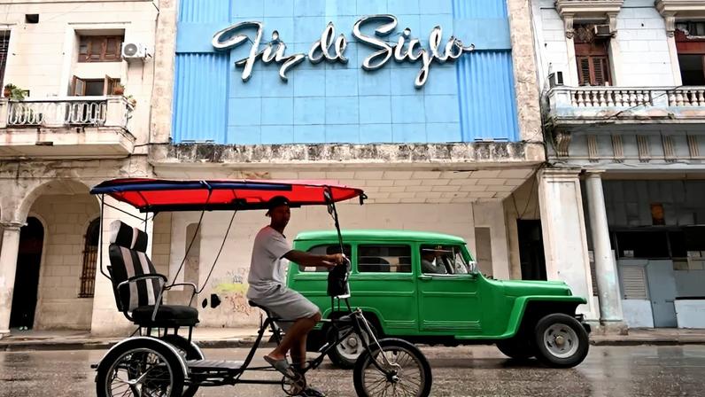 A bicycle taxi with a red roof and a green car drive along a street in Havana.
