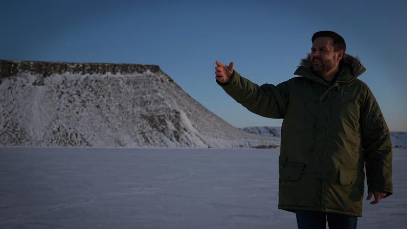 J.D. Vance dressed in a green parka gestures while standing in front of a snowy plateau.