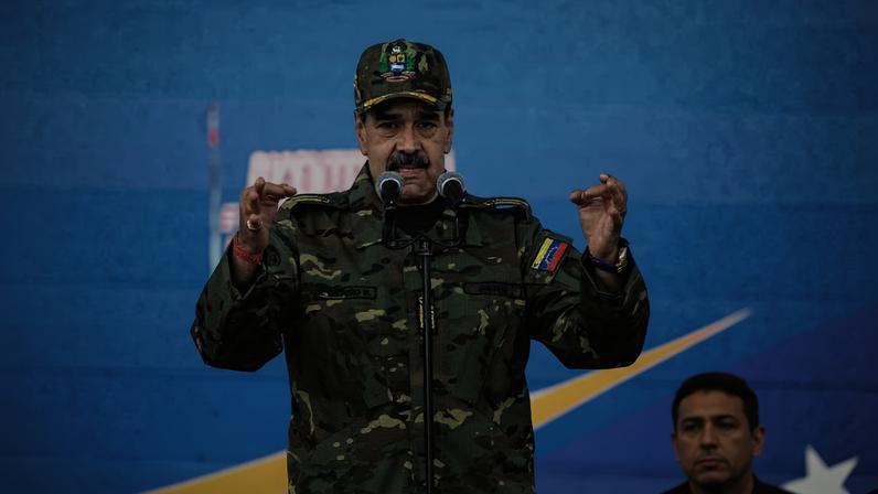 Nicolas Maduro is seen speaking at a military ceremony, wearing fatigues, with other military members and officials behind him.