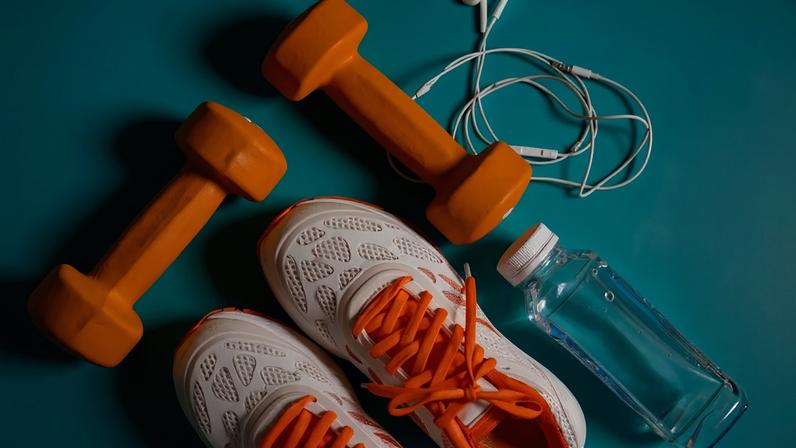 Imagem, vista de cima em fundo verde, de um par de tênis branco e laranja, uma garrafa d'água, um fone de ouvido e dois pesos pequenos para musculação.