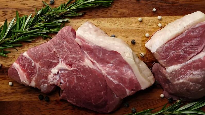 Imagem de dois pedaços de carne de porco em cima de uma tábua de madeira. Em volta, vê-se ramos de alecrim e bolinhas de pimenta do reino.