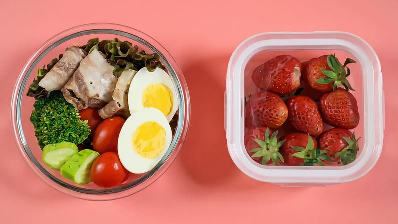 Imagem, em fundo rosa, de um pote de vidro contendo ovos e legumes. Ao lado, vê-se um pote de plástico contendo morangos.