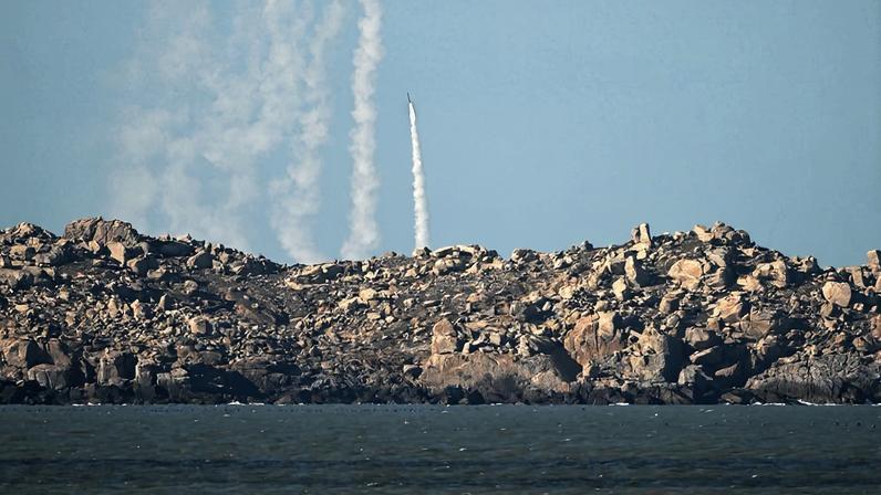 A rocket is seen from a distance as it launches against a blue sky from the rocky surface of an island, a plume of white smoke billowing from beneath it. Other white plumes are visible to the side.