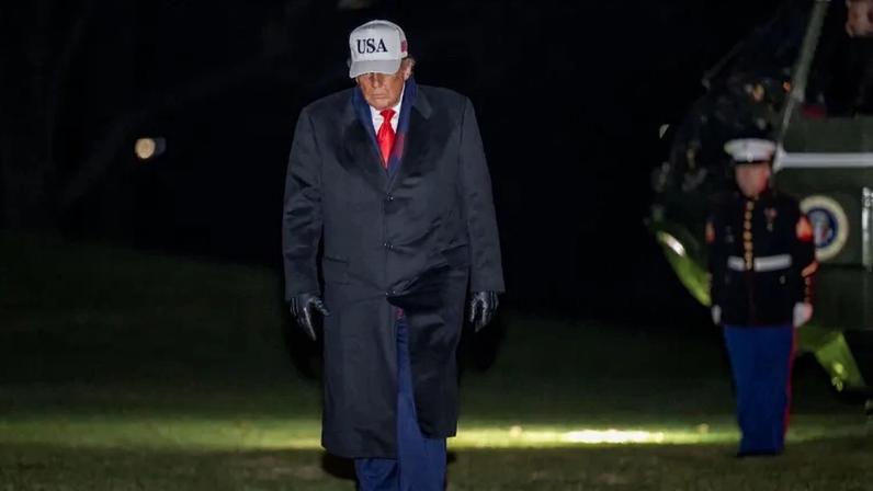 Donald Trump cuts an imposing figure against a dark nighttime background as he walks across a lawn with a parked helicopter and uniformed military officer standing in front of it. Trump wears a long black coat over a navy suit and red tie, along with a grey ballcap hat reading "USA." U.S. President Donald Trump walks from Marine One to the White House in Washington on Dec. 13, 2025.