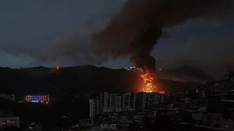 A plume of black smoke and orange fire billows from a sprawling midrise building complex. Dark shadows of mountains loom in the distance beneath a dim sky. A large building in the foothills of the mountains is lit up in the colors of the Venezuelan flag—yellow, blue, and red.