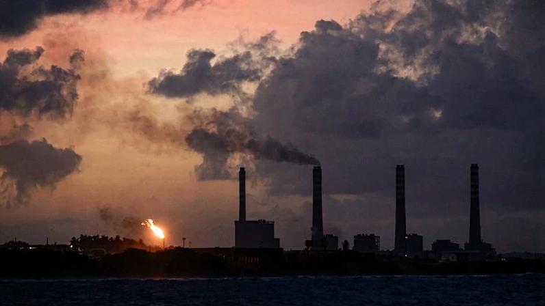 The dark silhouettes of oil refinery buildings are seen against a dusky sky.