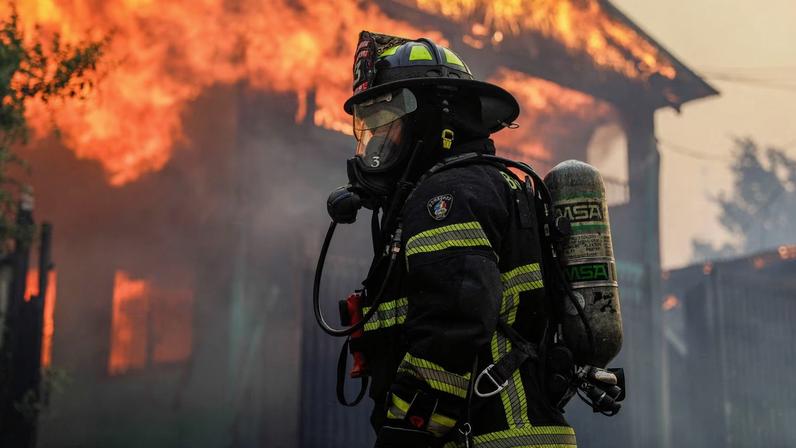 A firefighter stands in front of a burning building as fire and smoke rise from a forest fire in the Biobio region where, according to local media, multiple wildfires prompted emergency evacuations, in Concepcion, Chile, January 18, 2026. REUTERS/Juan Gonzalez