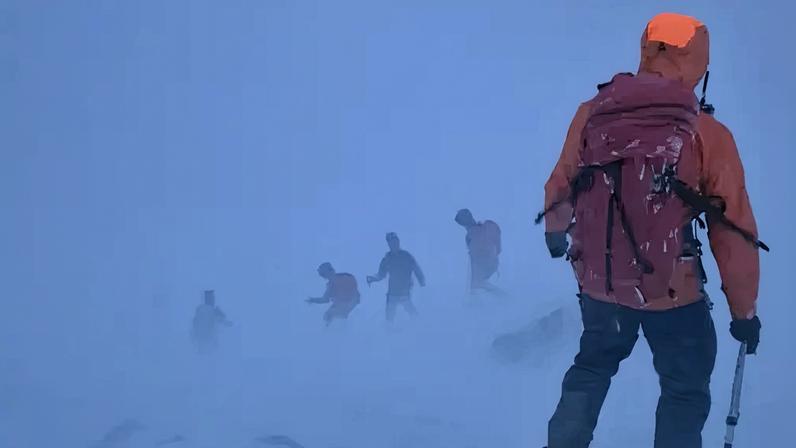 Glencoe MRT Glencoe MRT members in blizzard conditions. The volunteer in the foreground has a red jacket with the hood up and black waterproof trousers. In front of him are four figures walking through wind-blown snow.