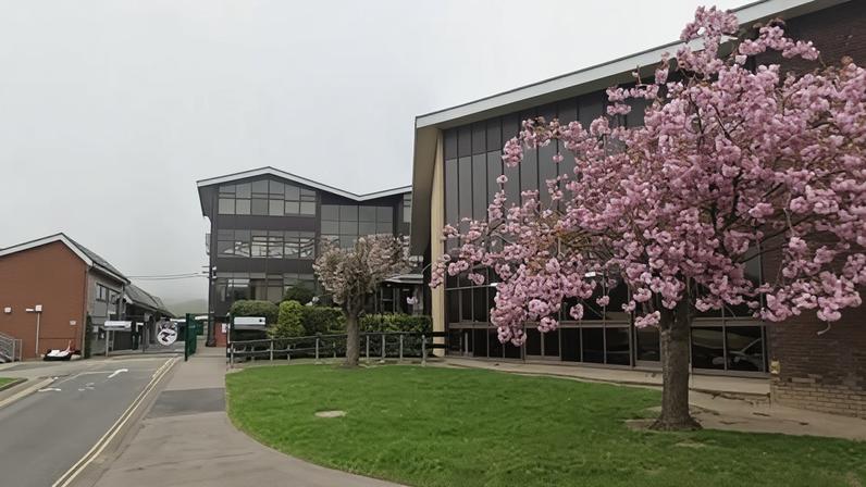 The outside of a school in Scarborough, with a road leading to the school buildings. To the right of the image is a green space with cherry blossom trees in full bloom.