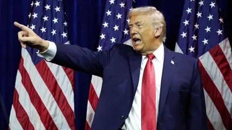 Getty Images US President Donald Trump, wearing a blue suit and red tie, points with his finger in front of an American flag.