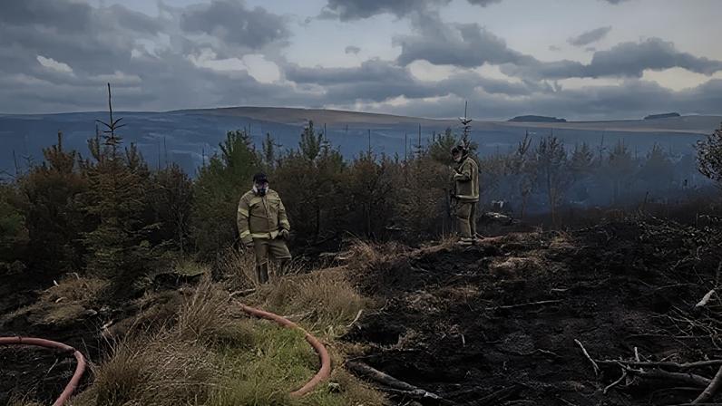 BBC Two firefighters with a large hose on Langdale Moor. They are facing a burned out patch of ground. There is smoke hanging in the air around them