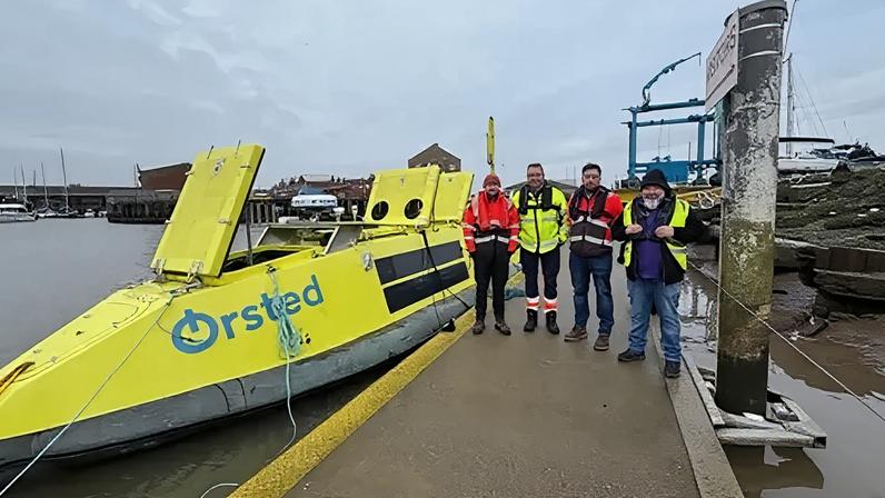 Four men in high-vis clothing stand on a gangway next to a fluorescent yellow boat, which has Orsted branding on the side. Grimsby Docks can be seen in the background.