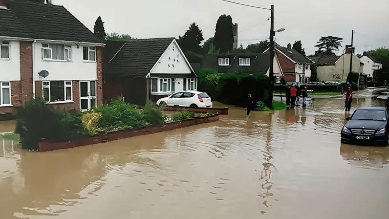 Gary Manning A residential street is heavily flooded, with muddy water covering the road and reaching up to the front gardens of houses. Several people stand on the higher, drier parts of the street observing the scene, while cars are partially submerged in the floodwater.