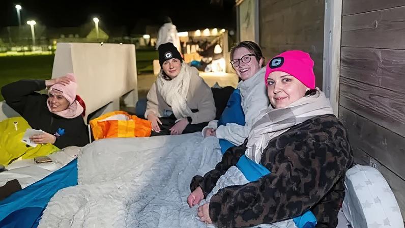 Skipton International Four women sitting on the ground outside wearing warm clothes, all in sleeping bags. It's around the evening and in the background you can see a sports field and other people in their sleeping bags and warm clothes. All of the women are looking at the camera smiling.