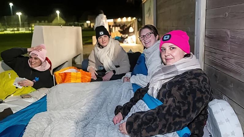 Skipton International Four women sitting on the ground outside wearing warm clothes, all in sleeping bags. It's around the evening and in the background you can see a sports field and other people in their sleeping bags and warm clothes. All of the women are looking at the camera smiling.