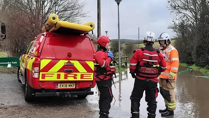Dorset Police Three firefighters are stood in conversation next to a patch of land flooded with water and a red van with the word 'Fire' printed on the back in large letters. The surroundings are rural, with several bare trees and green fields in the background. To the left of the van is a green sign that reads "Thornicombe Park".