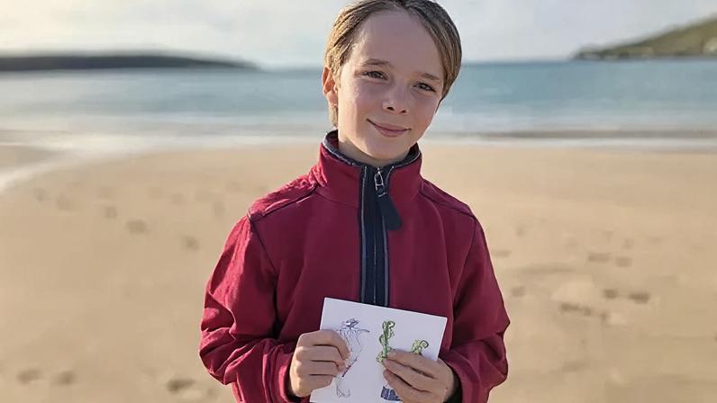 BBC A young boy with blond hair and a red fleece stands of a beach in the sunshine, hoping an illustrated greetings card