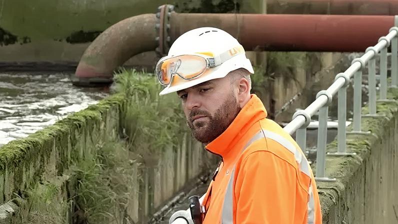 Ben Schofield/BBC An Anglian Water employee inspecting the sewage works at Bedford Water Recycling Centre. He is wearing a white hard hat, which has a pair of goggles laid over the top, and an orange high visibility jacket. He has a closely cropped beard and is looking down and to the left of the image. He is standing against a guard rail, beyond which is a body of water, concrete walls and in the distance behind him a large, industrial looking red pipe. The pipe runs from the top right of the image, behind the worker's head and then curves and disappears into the water. The concrete looks old and has moss and grass growing out of it at various points.
