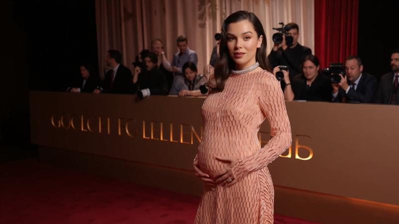 Hailee Steinfeld, in a peach gown with silver accents, poses on a red carpet while holding her pregnant belly.