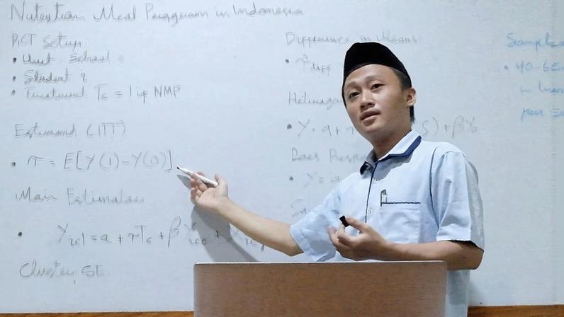 Munip Utama stands before a whiteboard, gesturing. A laptop sits open on the table in front of him.