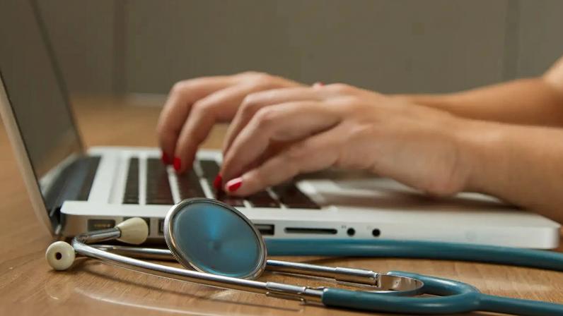 Médica digita no computador, tendo seu estetoscópio ao lado.