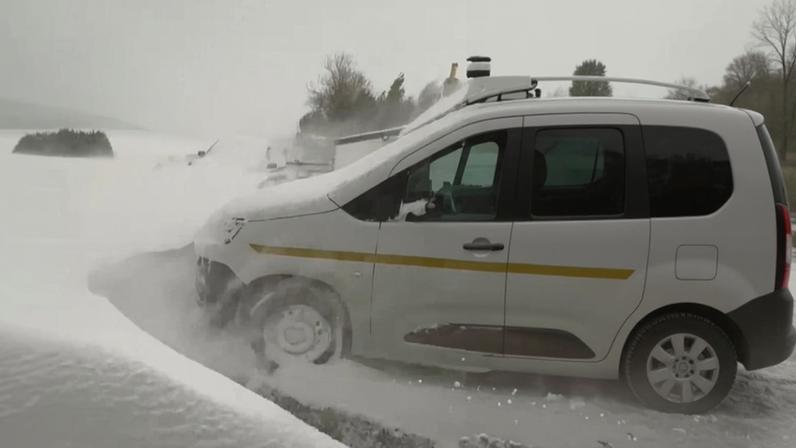The front of a parked van is surrounded by a high wall of snow, which is still falling.