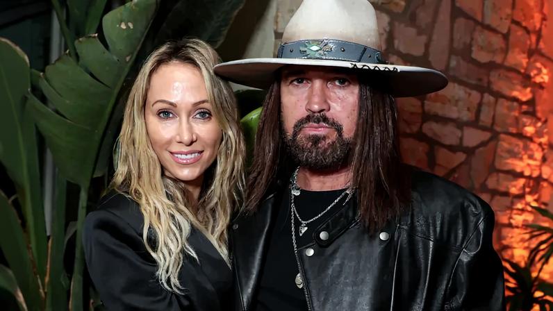 Tish Cyrus, in a black satin jacket, and Billy Ray Cyrus, in a black leather jacket and cowboy hat, pose for a photo.